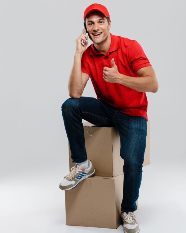 Young delivery man talking on cellphone while posing with cardboard boxes isolated over white wall